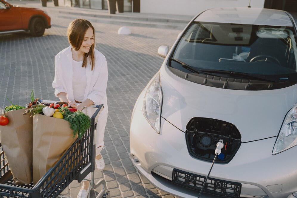 A woman in white attire stands next to a white electric car, with a grocery cart full of fresh produce beside her.