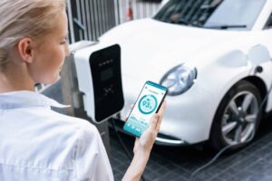 A person holds a smartphone displaying a 93% battery charge while an electric car is plugged into a charging station.