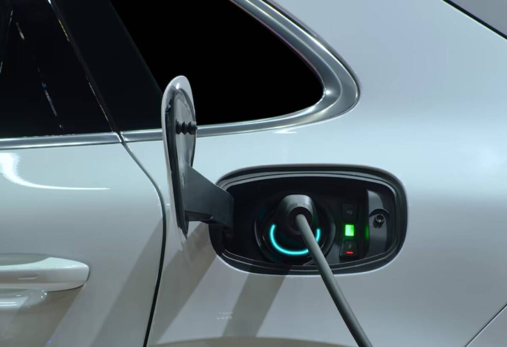 Close-up of a white electric vehicle's charging port with a plugged-in charger, showcasing charging indicators and a glossy finish.