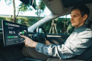 A person in a gray shirt points at a large touchscreen displaying a 60% charging status and time remaining of 6 hours 10 minutes in a car.
