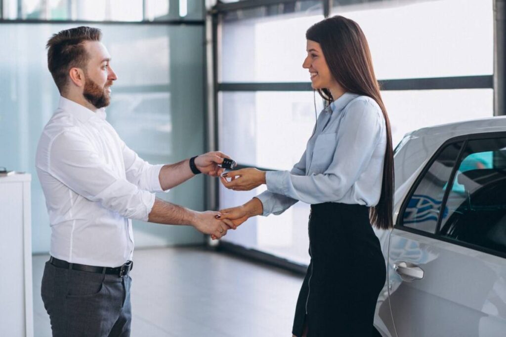 A man hands a car key to a woman in a car dealership, both smiling amidst a modern, well-lit showroom.