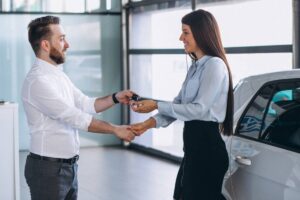 A man hands a car key to a woman in a car dealership, both smiling amidst a modern, well-lit showroom.