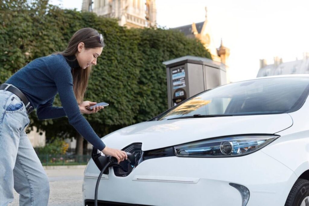 A person connects a charging cable to a white electric car, while holding a smartphone, at an outdoor charging station.