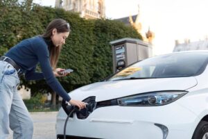 A person connects a charging cable to a white electric car, while holding a smartphone, at an outdoor charging station.