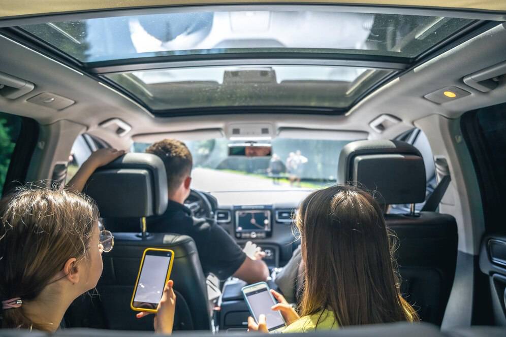 Two passengers in a car, engaged with their phones, while a driver focuses on the road ahead. Sunlight streams through the sunroof.