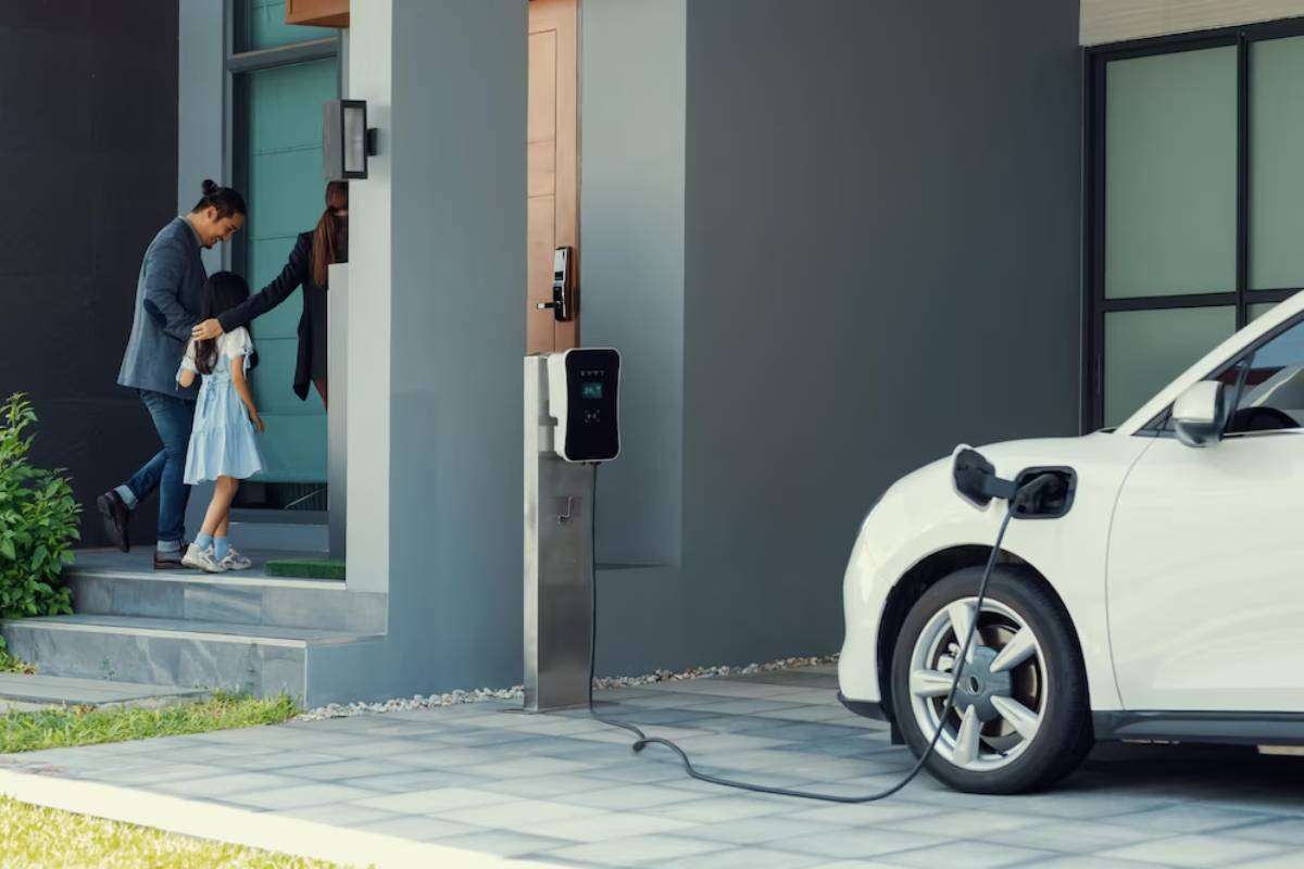 A white electric car is parked in a driveway, charging next to a modern home with family members entering the front door.