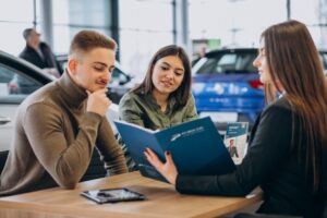 A group of people discusses details at a car dealership, reviewing a portfolio and brochures with various vehicles in the background.