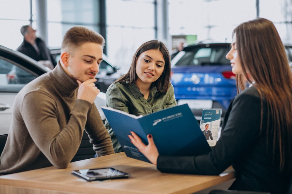 A group of people discusses details at a car dealership, reviewing a portfolio and brochures with various vehicles in the background.