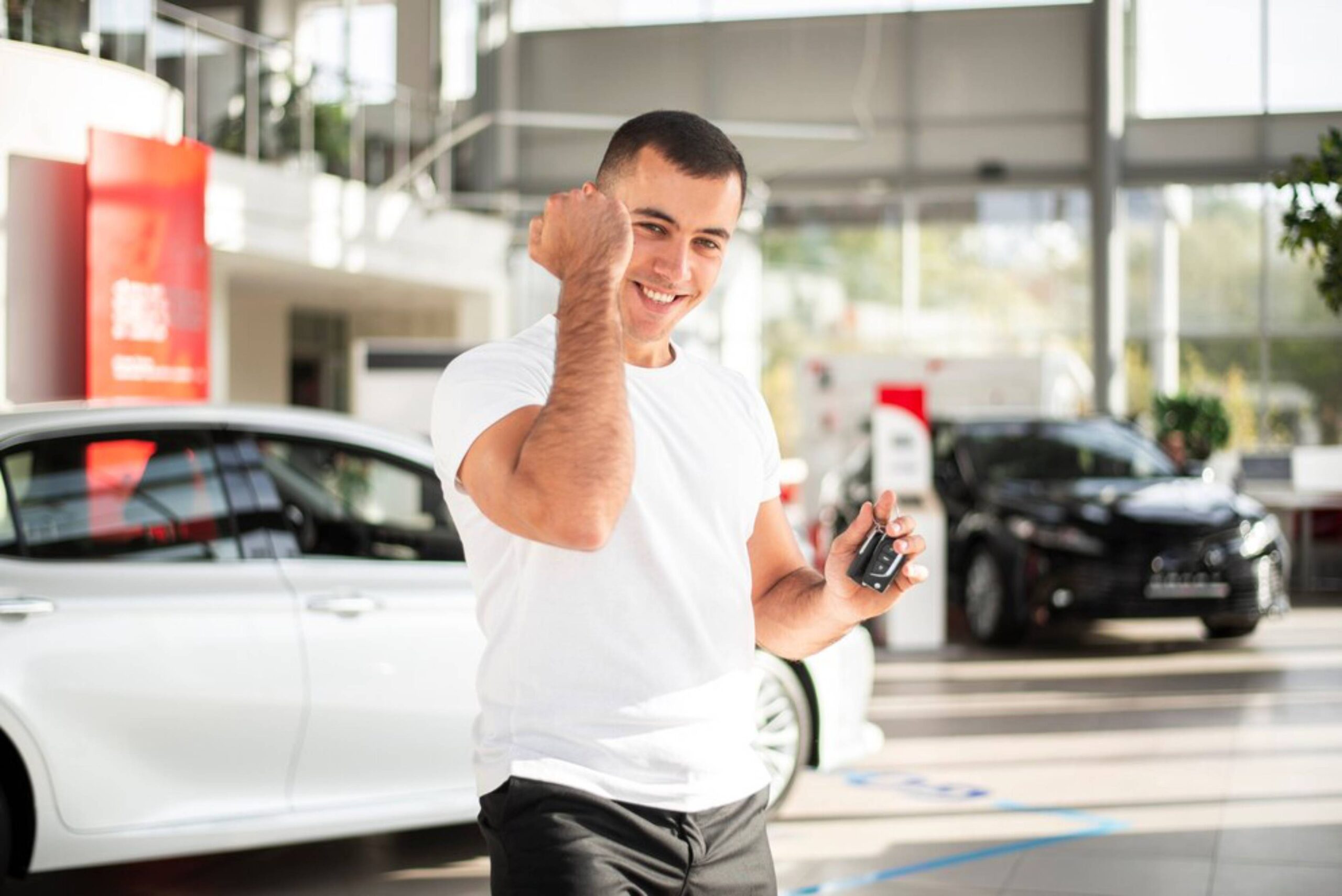 A man in a white t-shirt holds car keys and gestures, standing in a bright car dealership with modern vehicles in the background.