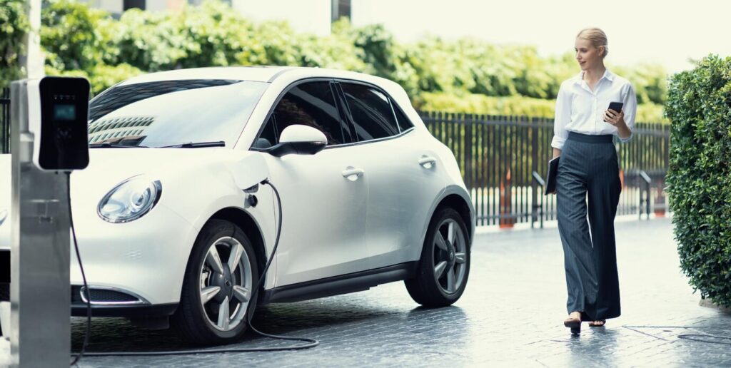 A stylish woman walks past a white electric car, plugged in at a charging station among greenery in an urban setting.