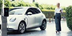 A stylish woman walks past a white electric car, plugged in at a charging station among greenery in an urban setting.